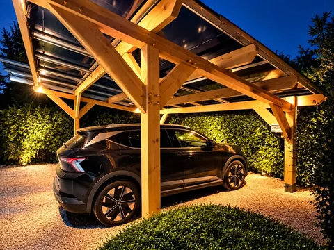 Carport en bois couvert de panneaux solaires, éclairé de nuit, abritant une voiture électrique noire
